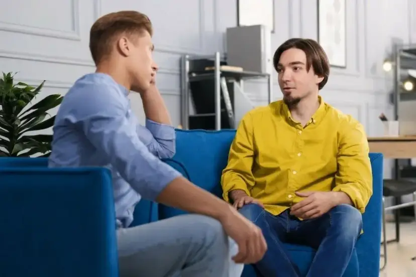 Dwóch mężczyzn siedzi na kanapie, rozmawiając. Jeden z nich opowiada o tym, jak przełamać barierę językową, szukając inspiracji w szczerej rozmowie.