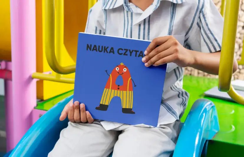 Chłopiec trzyma książkę "Nauka czytania dla 6 latka" z zabawną literką "A" na okładce.