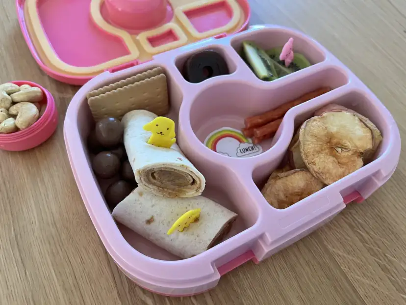 Różowy lunchbox z pysznym, zdrowym śniadaniem do szkoły: wrap z kurczakiem, suszone jabłka, kiwi, orzechy i czekoladowe kuleczki.
