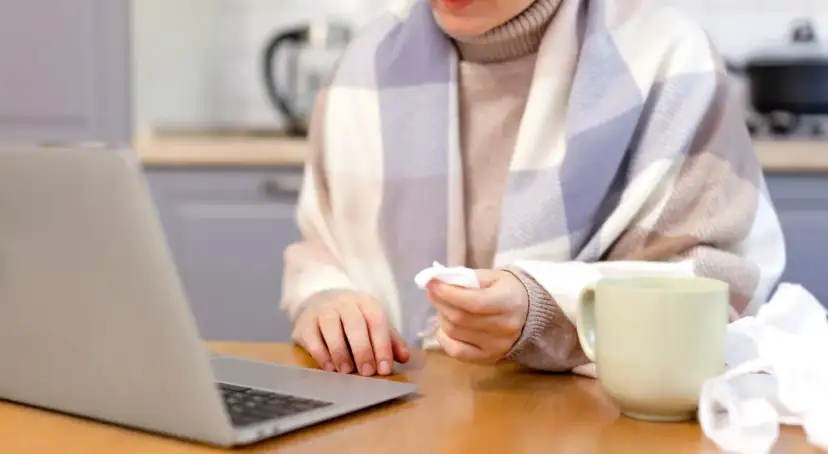 Osoba owinięta kocem, z chusteczką w dłoni, pracuje przy laptopie. Obok kubek i zużyte chusteczki. Może to być sytuacja związana z art. 148 kp.