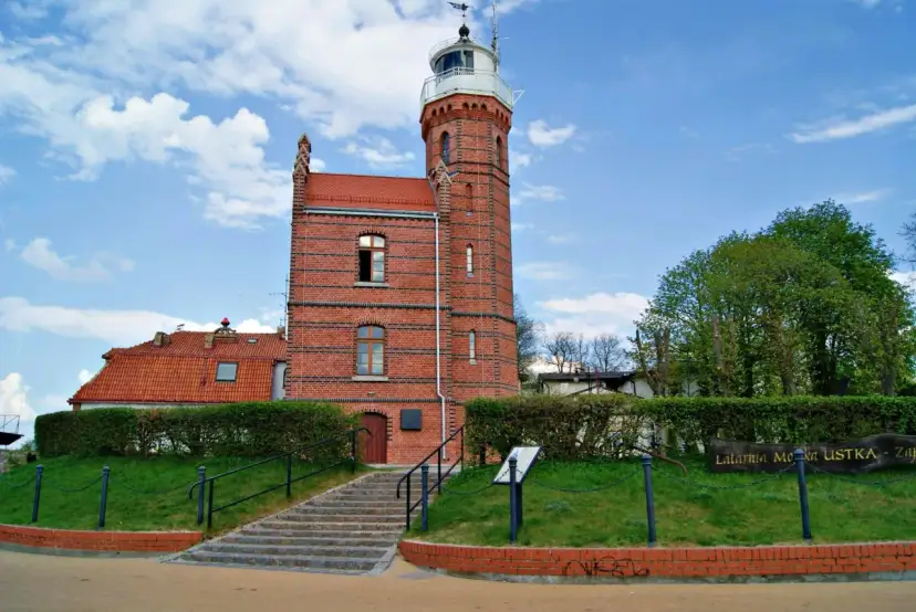 Ceglana latarnia morska w Ustce z czerwoną dachówką i zielonymi żywopłotami.