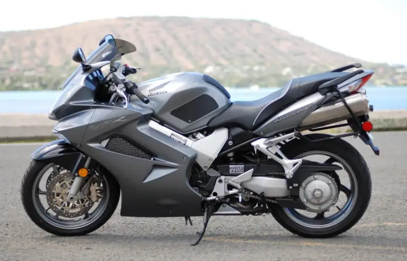 Szary motocykl Honda VFR 800 z widokiem na wodę i wzgórze w tle.