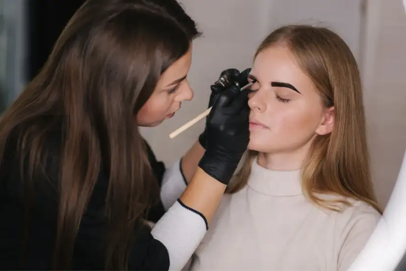 Stylistka w czarnych rękawiczkach precyzyjnie maluje brwi klientce. Czy to henna czy farbka, efekt będzie zachwycający.