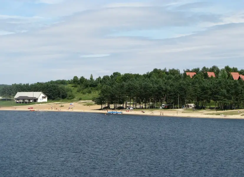 Plaża nad jeziorem Chańcza tętni życiem. Ludzie odpoczywają na piasku, kąpią się w wodzie, a w tle widać domki i las.