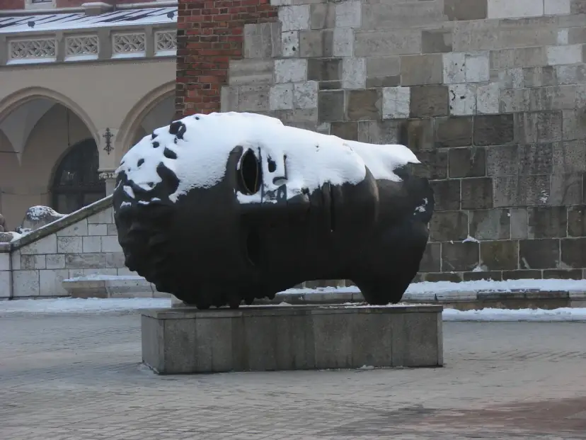 Duża, ciemna rzeźba głowy pokryta śniegiem, na tle kamiennej ściany i arkadowego budynku.