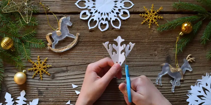 Ręce tnące papierową śnieżynkę na drewnianym tle z ozdobami świątecznymi.