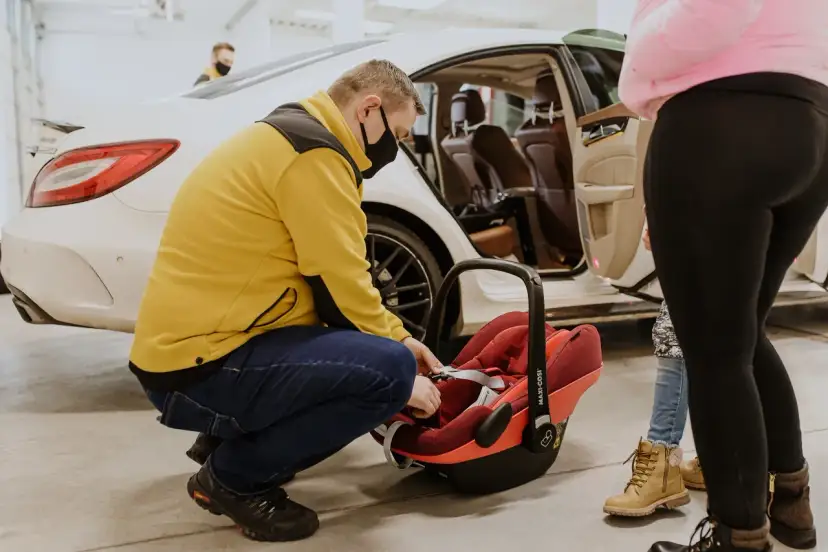 Mężczyzna w żółtej kurtce zapina pasy w czerwonej wkładce do nosidełka Maxi-Cosi, przygotowując ją do podróży samochodem.