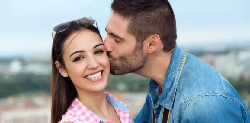 Całuje ją w policzek, jak pisze zakochany facet. Uśmiechnięta dziewczyna w kratkę i chłopak w dżinsowej kurtce.