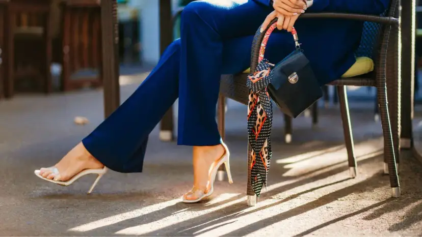 Kobieta w granatowym kombinezonie, białych sandałach na obcasie i z czarną torebką.