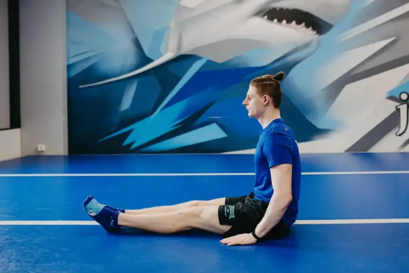 Mężczyzna w niebieskiej koszulce i czarnych spodenkach siedzi na niebieskiej macie z wyprostowanymi nogami.