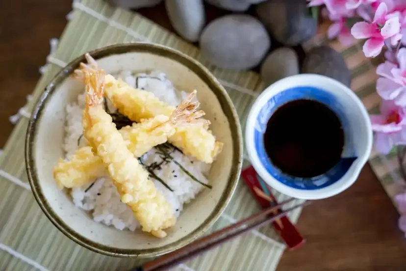 Chrupiące krewetki w cieście tempura na ryżu, obok miseczka z ciemnym sosem do tempury.