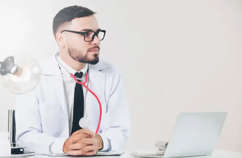 Lekarz medycyny pracy w okularach, z czerwonym stetoskopem, siedzi przy biurku z laptopem. Jakie badania wykonuje?