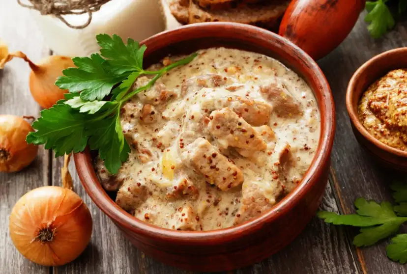 Gulasz wieprzowy w kremowym sosie musztardowym, ozdobiony natką pietruszki, obok cebuli i chleba.