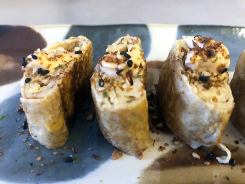 Trzy kawałki sushi w omlecie, polane majonezem i posypane czarnym sezamem.