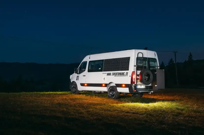 Biały kamper Renault Master z kołem zapasowym z tyłu, zaparkowany na łące nocą.