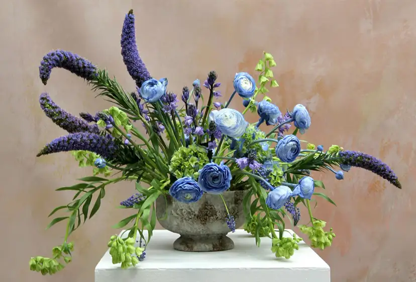 Kompozycja kwiatowa w stylu dekoracyjnym we florystyce, z niebieskimi różami, fioletowymi kłosami i zielonymi akcentami.