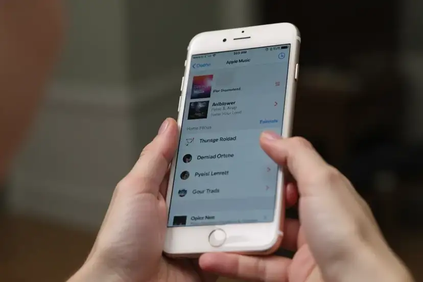 Ręce trzymają iPhone'a z otwartą aplikacją Apple Music. Widoczna lista utworów, która może pomóc w nauce, jak pobrać muzykę na iPhone'a.