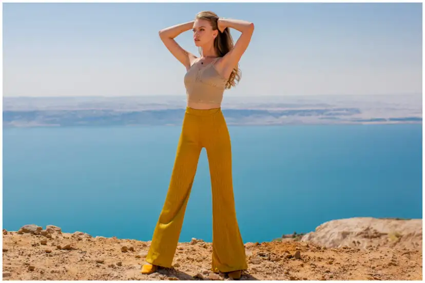 Kobieta w musztardowych spodniach dzwonach i beżowym topie na tle błękitnego jeziora.