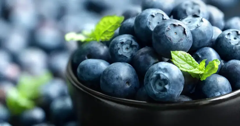 Miseczka pełna jagód, które są dobre na wzrok i serce. W tle widać więcej owoców i listki mięty.