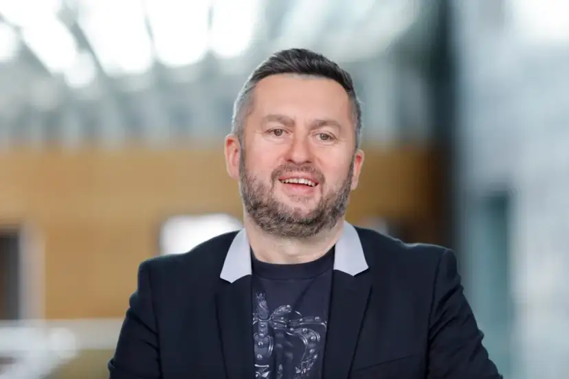 Uśmiechnięty mężczyzna w czarnej marynarce i t-shircie z nadrukiem. Ciekawe, ile zarabia Norbi w Kole Fortuny?