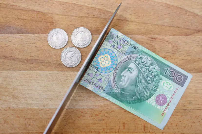 Nóż przecina banknot 100 zł, obok leżą trzy monety 1 zł.