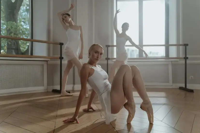 Trzy baletnice w białych strojach ćwiczą podnoszenia w tańcu. Jedna siedzi na podłodze, dwie inne stoją w pozycjach.