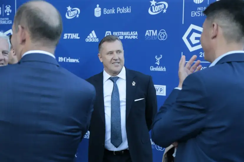 Mężczyzna w garniturze uśmiecha się, stojąc na tle niebieskiej ściany z logotypami sponsorów, być może rozmawiając o tym, ile dostają kluby ekstraklasy.