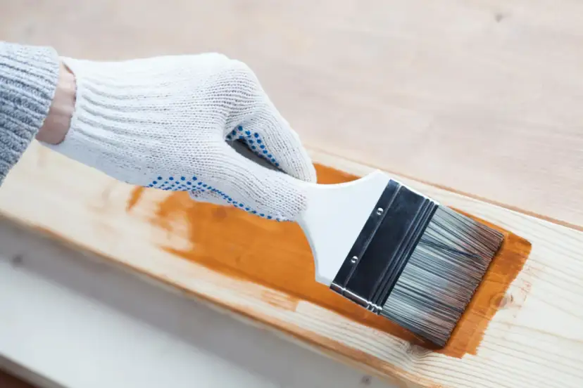 Ręka w białej rękawicy nakłada pędzlem brązowy preparat na drewno. Czy to lakier, czy lakierobejca?