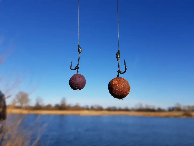 Dwa haczyki wędkarskie z kulkami proteinowymi na tle błękitnego nieba i wody.