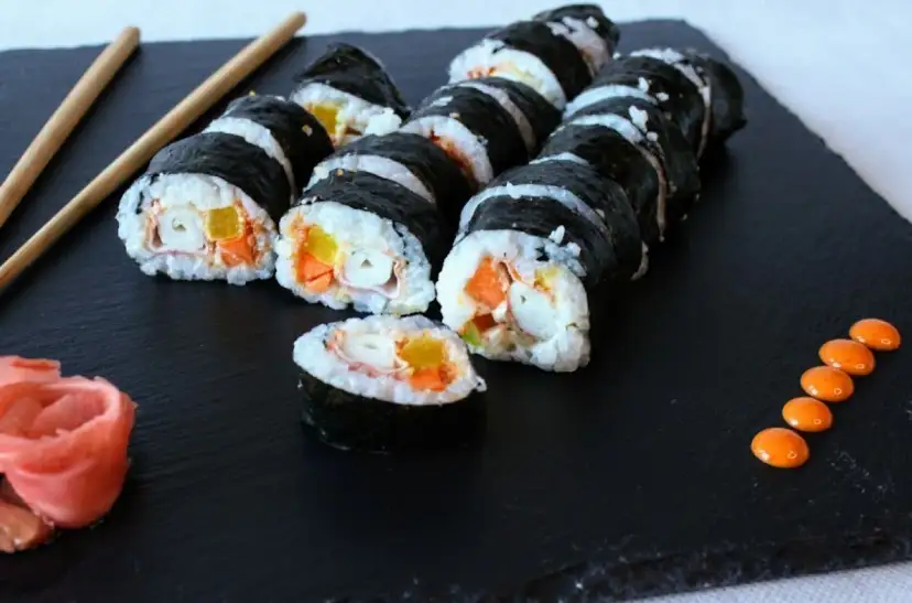 Roladki sushi z surimi, ryżem i warzywami na czarnej desce, obok pałeczki i różowy imbir.