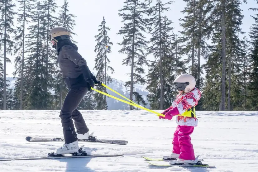 Dziecko w kombinezonie narciarskim uczy się jeździć na nartach, trzymając żółtą linkę.