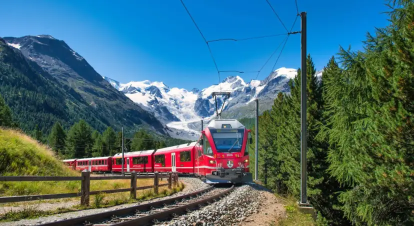 Czerwony pociąg przemierza malowniczą Szwajcarię, mijając zielone lasy i ośnieżone szczyty gór.