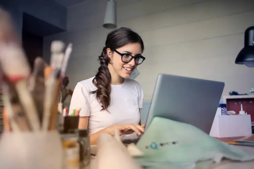 Uśmiechnięta kobieta w okularach pracuje przy laptopie, realizując swoją pracę dodatkową z domu.
