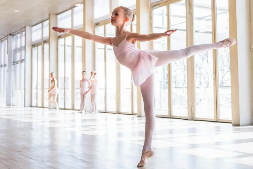 Młoda baletnica w różowej sukience wykonuje eleganckie pozycje baletowe. W tle inne tancerki.