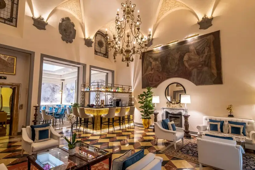 Elegant hotel lounge with a bar, fireplace, ornate chandelier, and artwork.
