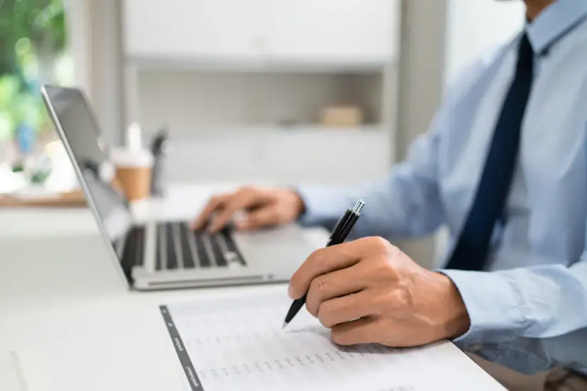 Mężczyzna w biurze pracuje przy laptopie i wypełnia dokumenty. Dowiaduje się, jak złożyć wniosek o wakacje kredytowe.