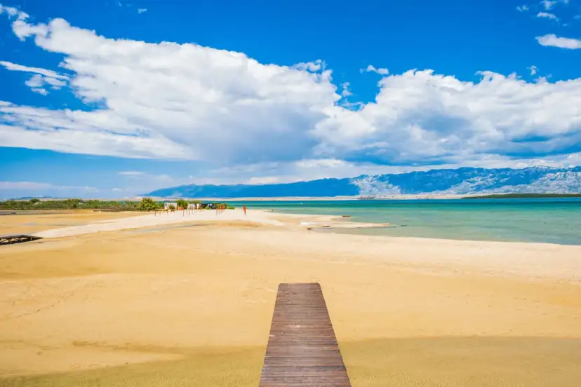 Drewniany pomost prowadzi na piaszczystą plażę Ninskiej laguny. W tle turkusowe morze i góry.