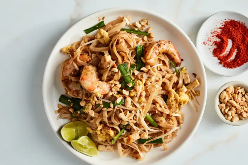 Talerz z Pad Thai z krewetkami, orzeszkami ziemnymi, jajkiem i limonką, obok miseczki z orzeszkami i papryką.