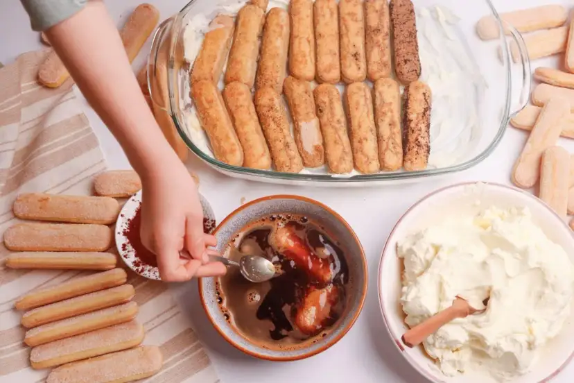 Dziecko zanurza biszkopty w kawie, przygotowując warstwy do tiramisu. Obok krem i kakao.