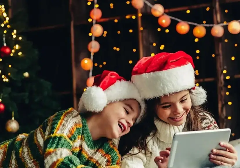 Dos niños con gorros de Santa Claus disfrutan de la tablet, felices cuando dan vacaciones de Navidad, rodeados de luces festivas y un árbol.