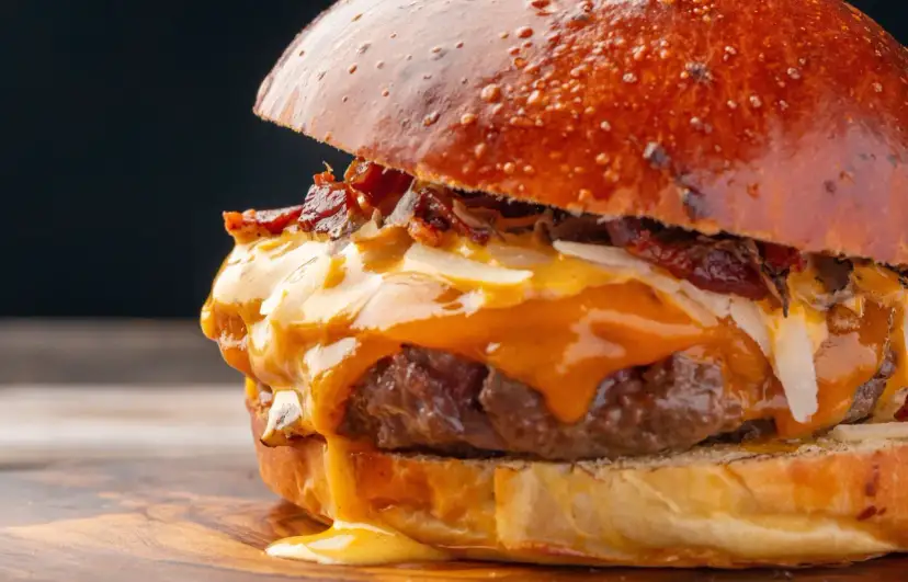 Soczysty burger z rozpływającym się serem, chrupiącym boczkiem i warstwami dodatków na drewnianej desce.