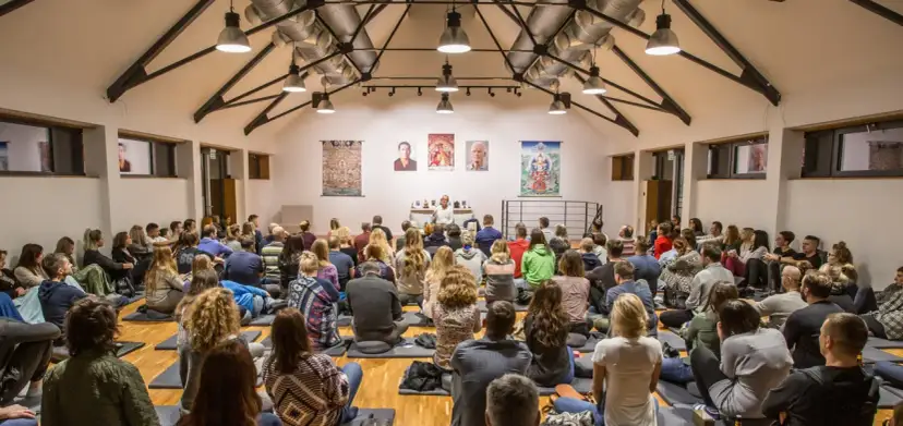 Wielka sala wypełniona ludźmi siedzącymi na podłodze, słuchającymi wykładu. Na ścianie wiszą buddyjskie obrazy, sugerując, że to miejsce, gdzie jest buddyzm.