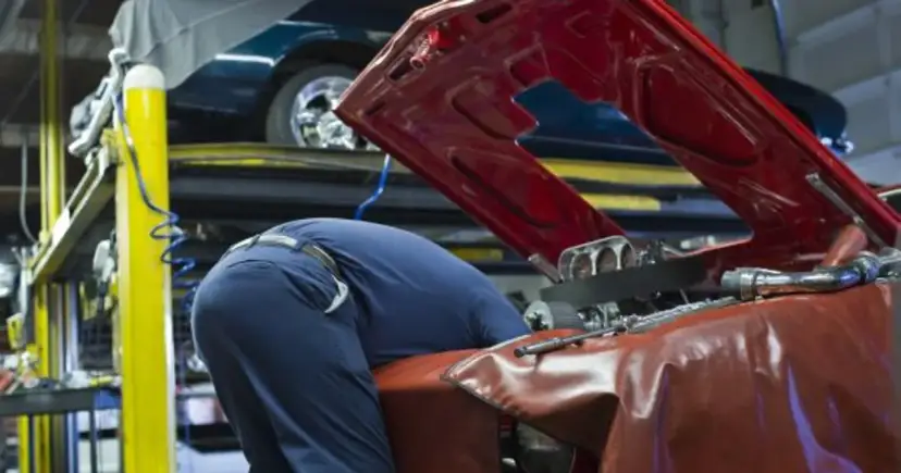 Mechanik w niebieskim kombinezonie pochyla się nad otwartą maską czerwonego samochodu.