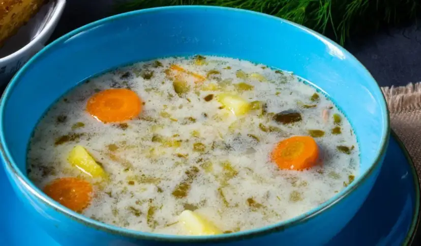 Zupa ogórkowa w niebieskiej misce z kawałkami marchewki i ziemniaków.