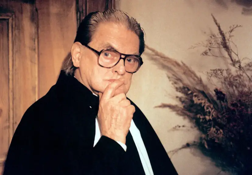 Rafał Dajbor w okularach, z dłonią na brodzie, w czarnym stroju, na tle drewnianej boazerii i bukietu suszonych traw.