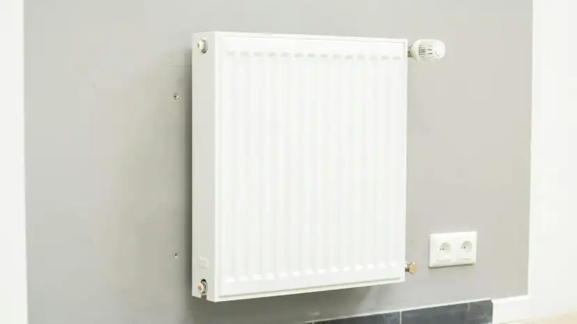 Biały grzejnik z termostatem na szarej ścianie. Dowiedz się, jak wymienić grzejnik w sezonie grzewczym, aby zapewnić ciepło w domu.