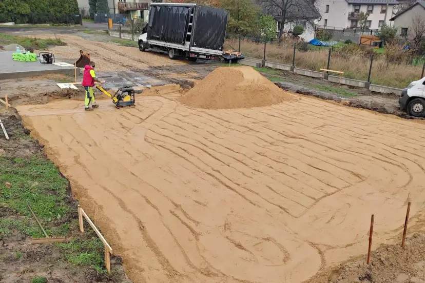 Pracownik zagęszcza piasek zagęszczarką. To kluczowy etap wymiany gruntu pod płytę fundamentową.