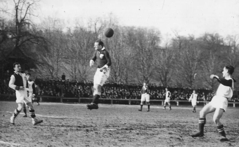 Piłkarz wyskakuje do główki, by trafić w piłkę. Na boisku widać innych graczy i trybuny pełne widzów.