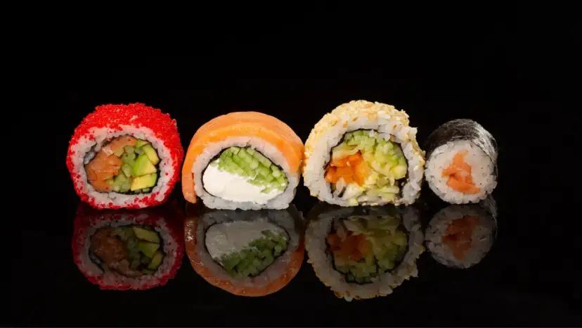 Różne rodzaje sushi: rolka z łososiem i awokado w ikrze, z łososiem i serkiem, z sezamem i warzywami, oraz klasyczne maki z łososiem.