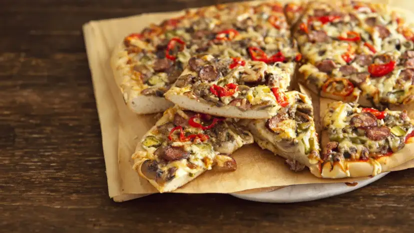 Grube ciasto na pizze z kiełbasą, pieczarkami i papryką, podane na papierze do pieczenia.